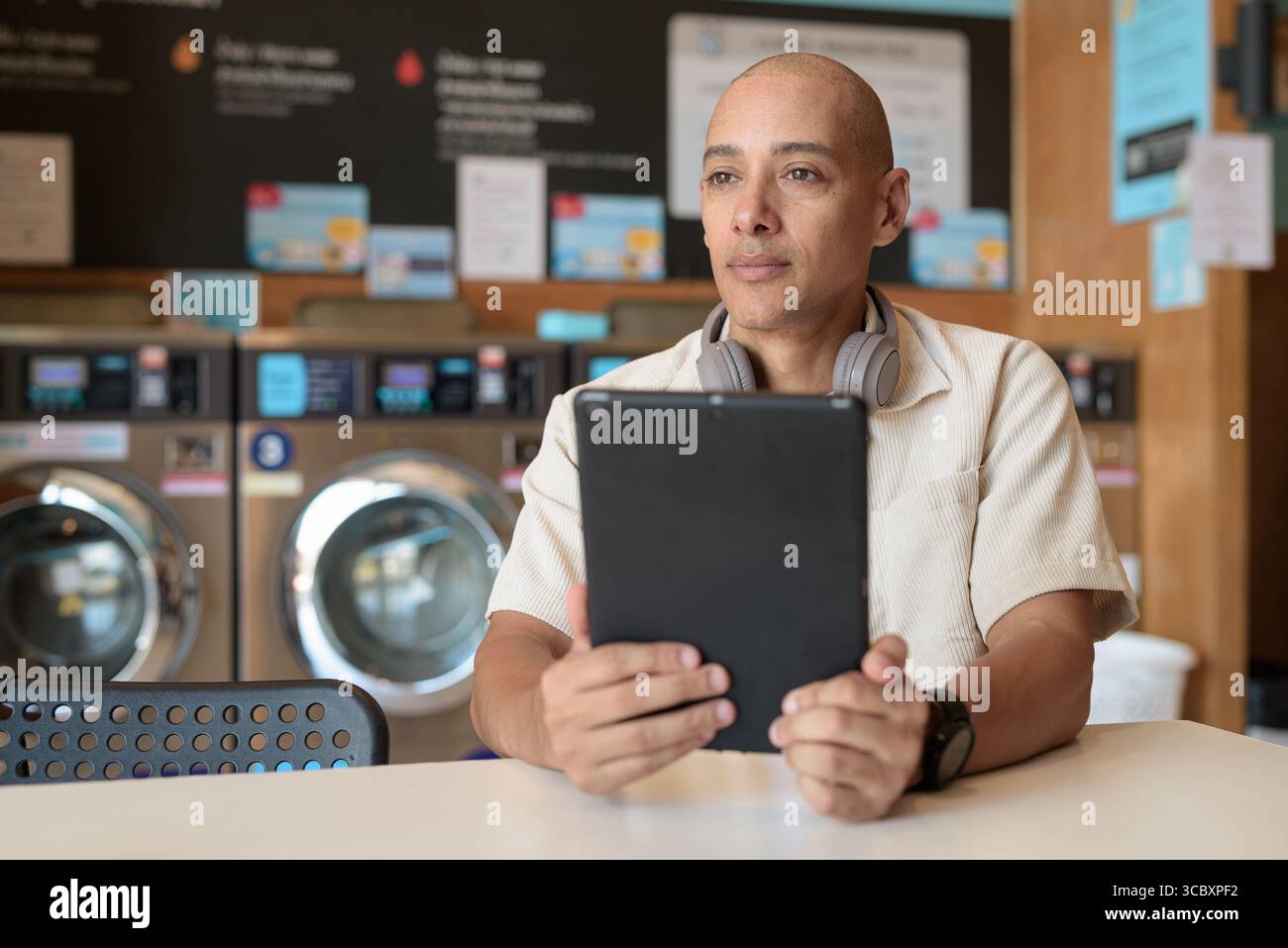 Kahler hispanischer Mann in lässiger Kleidung, der in einer Wäscherei sitzt und ein digitales Tablet benutzt. Urbanes Alltagskonzept mit Waschmaschinen im Hintergrund Stockfoto