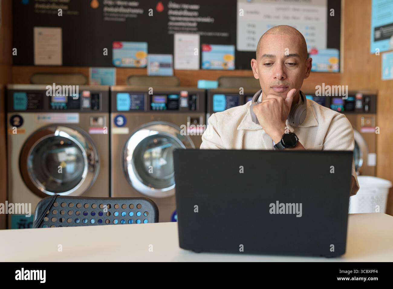 Kahler hispanischer Mann in legeren Kleidern, der in einem Wäscheshop sitzt und einen Laptop benutzt. Urbanes Alltagskonzept mit Waschmaschinen im Hintergrund Stockfoto