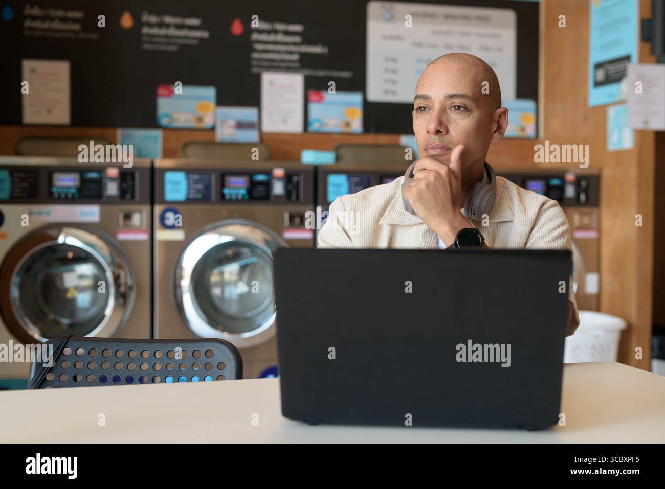 Kahler hispanischer Mann in legeren Kleidern, der in einem Wäscheshop sitzt und einen Laptop benutzt. Urbanes Alltagskonzept mit Waschmaschinen im Hintergrund Stockfoto