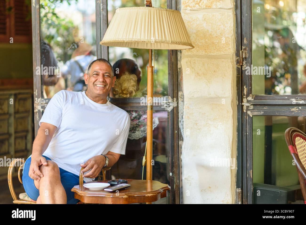 Ein lächelnder Mann sitzt beiläufig an einer Lampe in einem Café im Freien, sonnt sich im warmen Sonnenlicht und genießt einen entspannten Moment, umgeben von einer freundlichen Atmosphäre und Leuten, die sich in der Nähe unterhalten. Stockfoto