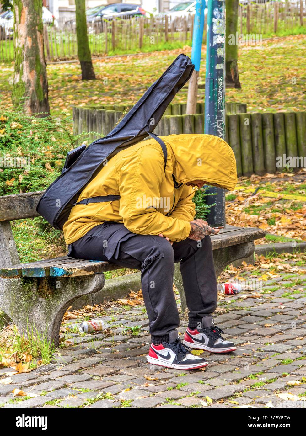 Junger Mann saß auf der Parkbank mit Gitarre auf dem Rücken und schaute auf das Smartphone - Brüssel, Belgien. Stockfoto