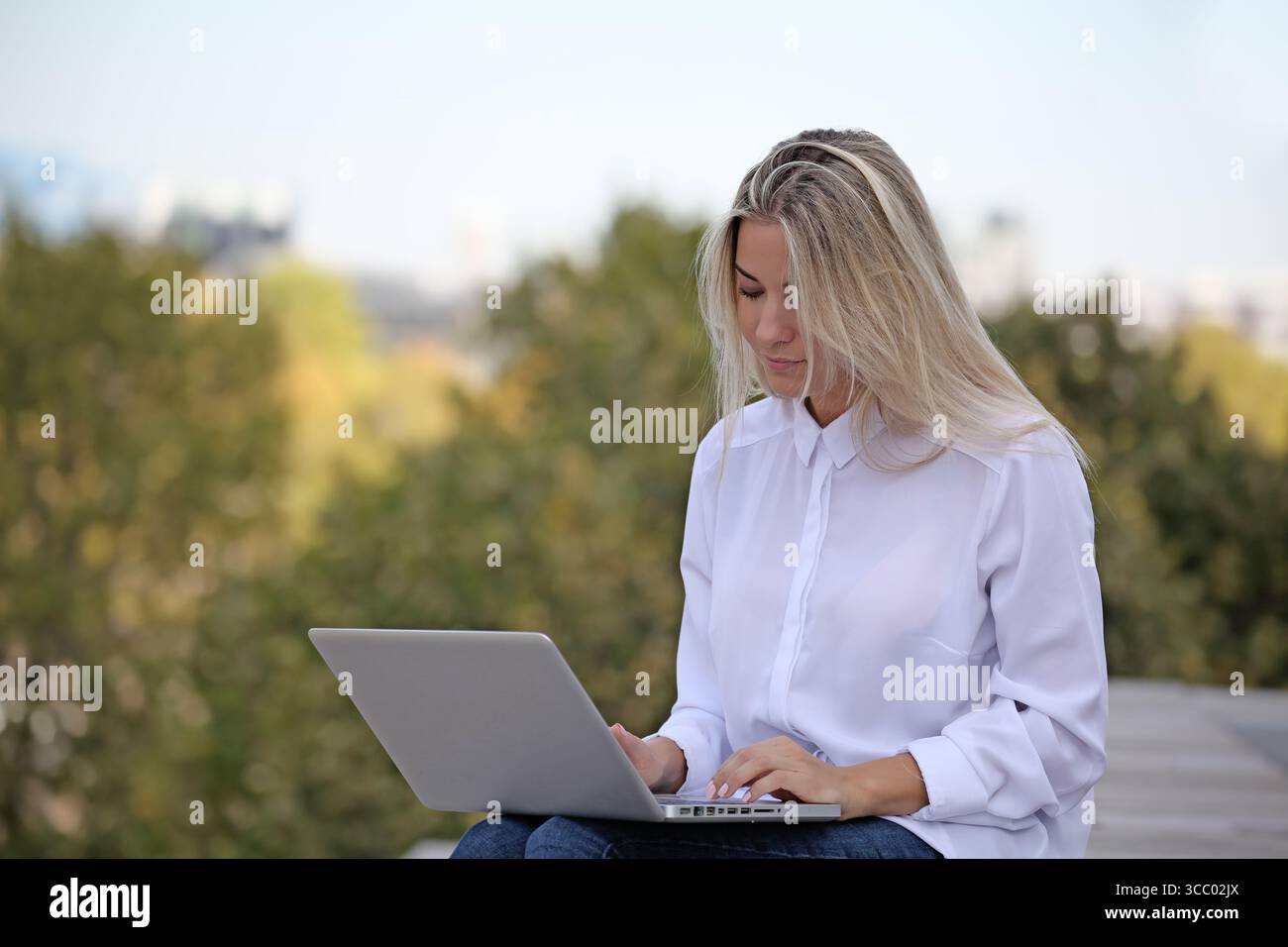 JUNGE BLONDE FRAU, DIE DRAUSSEN AUF IHREM LAPTOP ARBEITET Stockfoto JUNGE BLONDE FRAU, DIE DRAUSSEN AUF IHREM LAPTOP ARBEITET Stockfoto
