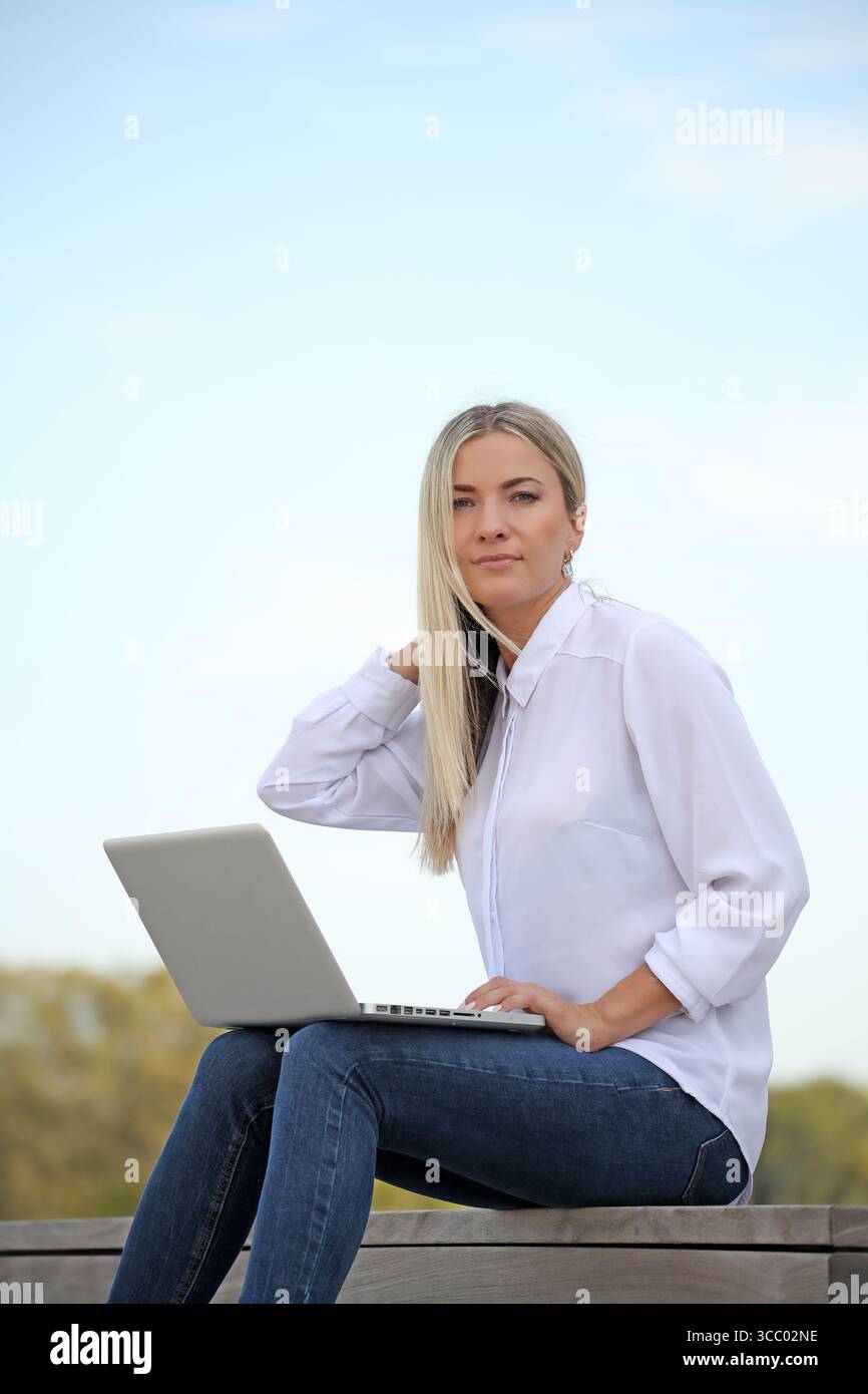 JUNGE BLONDE FRAU, DIE DRAUSSEN AUF IHREM LAPTOP ARBEITET Stockfoto JUNGE BLONDE FRAU, DIE DRAUSSEN AUF IHREM LAPTOP ARBEITET Stockfoto