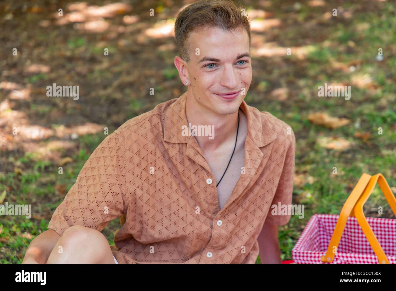 Porträt eines jungen Mannes, der beim Picknick in einem Park lächelt und bequem auf einer Decke sitzt, mit einem Picknickkorb gefüllt mit köstlichem Essen und Stockfoto