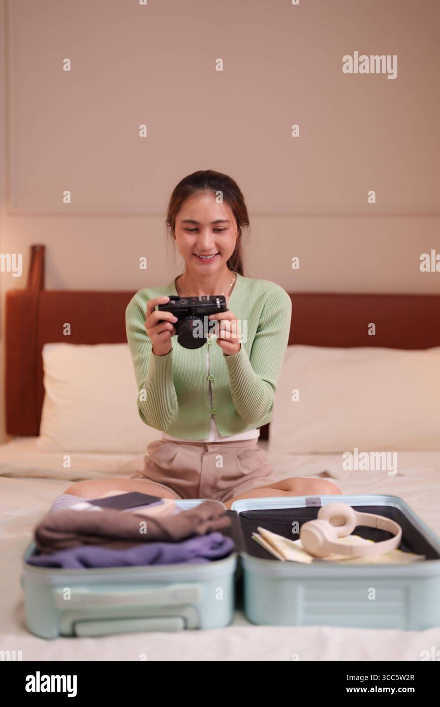 Lächelnde asiatische Frau, die im Hotel Koffer packt. Momente mit einer Vintage-Kamera festhalten. Bereit für aufregende Reiseabenteuer. Stockfoto