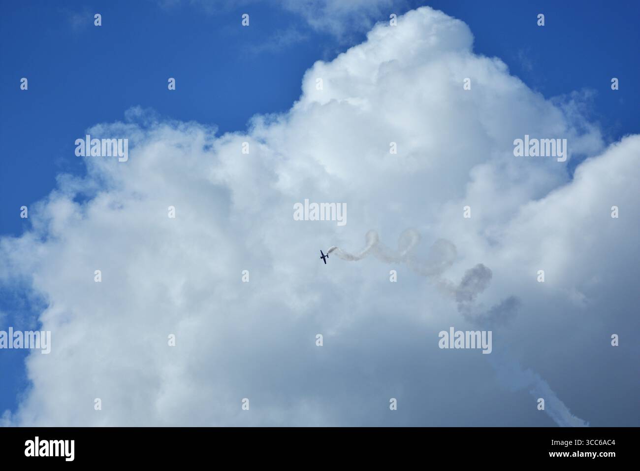 Kunstflugzeug, das während der Flugshow Spiralmanöver mit Rauchspur gegen hellblauen Himmel und aufragende weiße Wolken durchführt. Stockfoto