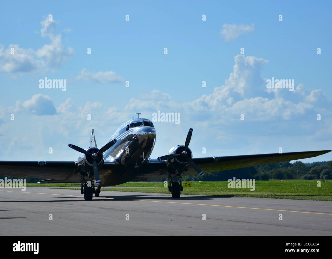 Glänzendes silbernes DC-3-Oldtimer-Flugzeug auf der Landebahn, Vorderansicht mit drehenden Doppelpropellern, heller sonniger Tag mit blauem Himmel und verstreuten Wolken. Stockfoto