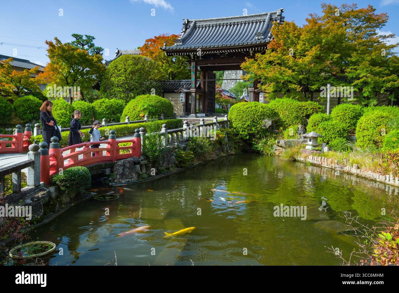 Ein ruhiger japanischer Garten mit einer Familie auf einer roten Brücke, üppigem Grün und einem friedlichen Teich mit Koi-Fischen. Die traditionelle Architektur verstärkt die Stockfoto