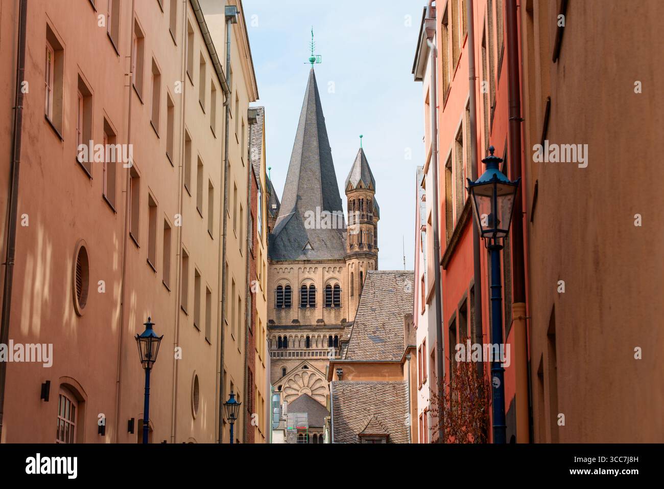 Köln, Deutschland - 15. Juni 2025: Schmaler Straßenblick in Köln umrahmt von bunten Gebäuden, mit einem markanten Kirchturm im Hintergrund und Stockfoto