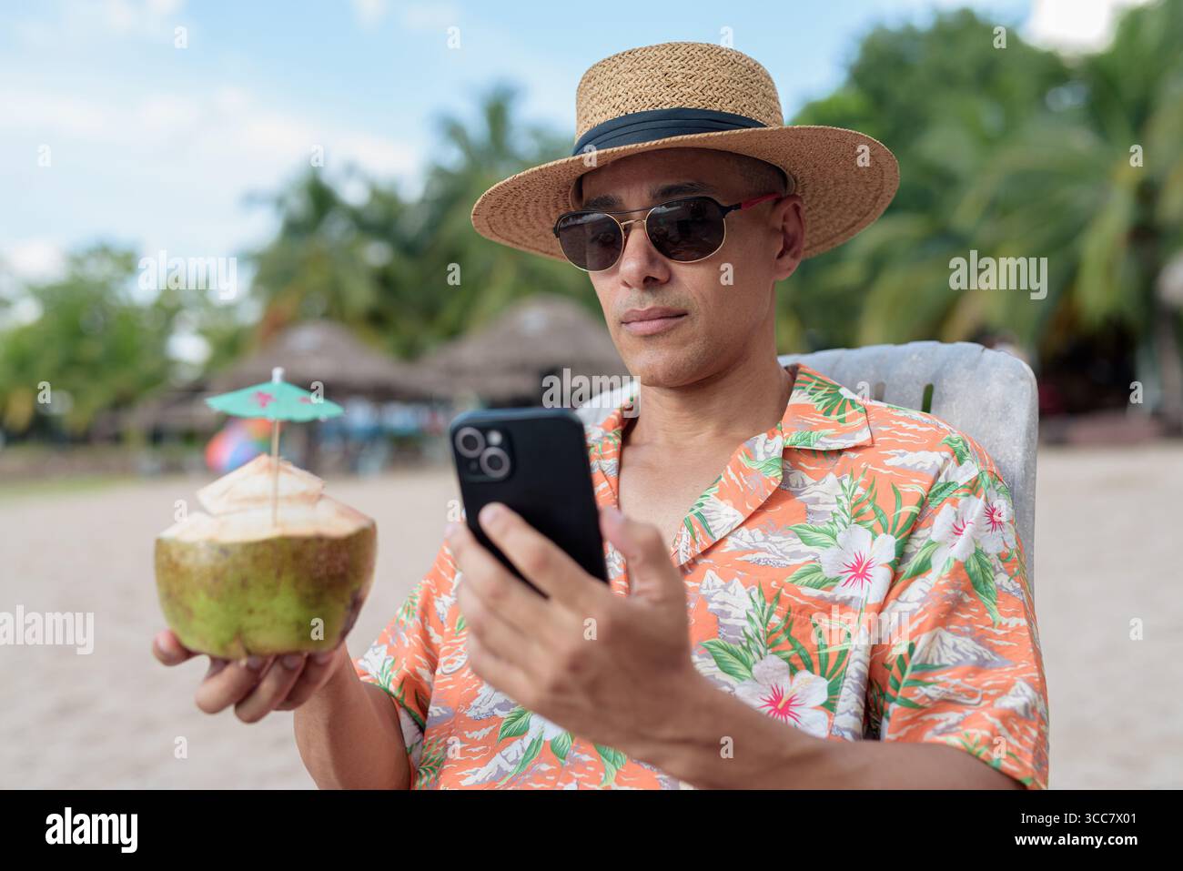 Hispanischer Mann mit Panamahut und hawaiianischem Hemd, gekleidet im lässigen Sommerurlaub-Stil. Er posiert am Strand und zeigt eine entspannte Urlaubsatmosphäre Stockfoto