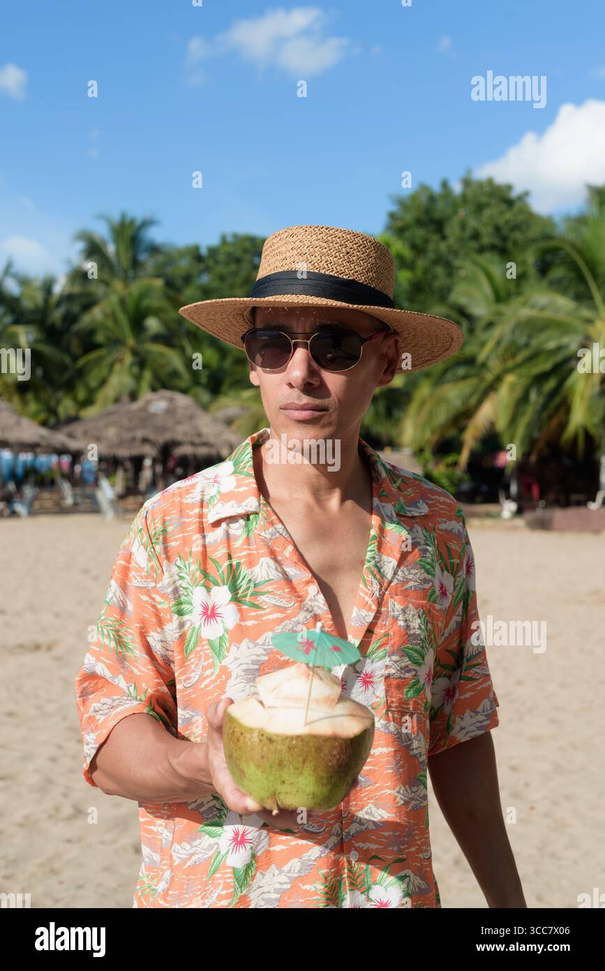 Hispanischer Mann mit Panamahut und hawaiianischem Hemd, gekleidet im lässigen Sommerurlaub-Stil. Er posiert am Strand und zeigt eine entspannte Urlaubsatmosphäre Stockfoto