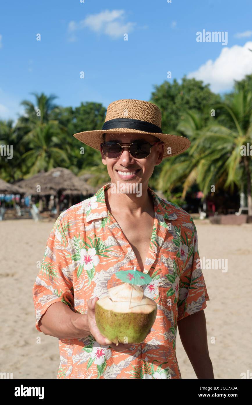 Hispanischer Mann mit Panamahut und hawaiianischem Hemd, gekleidet im lässigen Sommerurlaub-Stil. Er posiert am Strand und zeigt eine entspannte Urlaubsatmosphäre Stockfoto