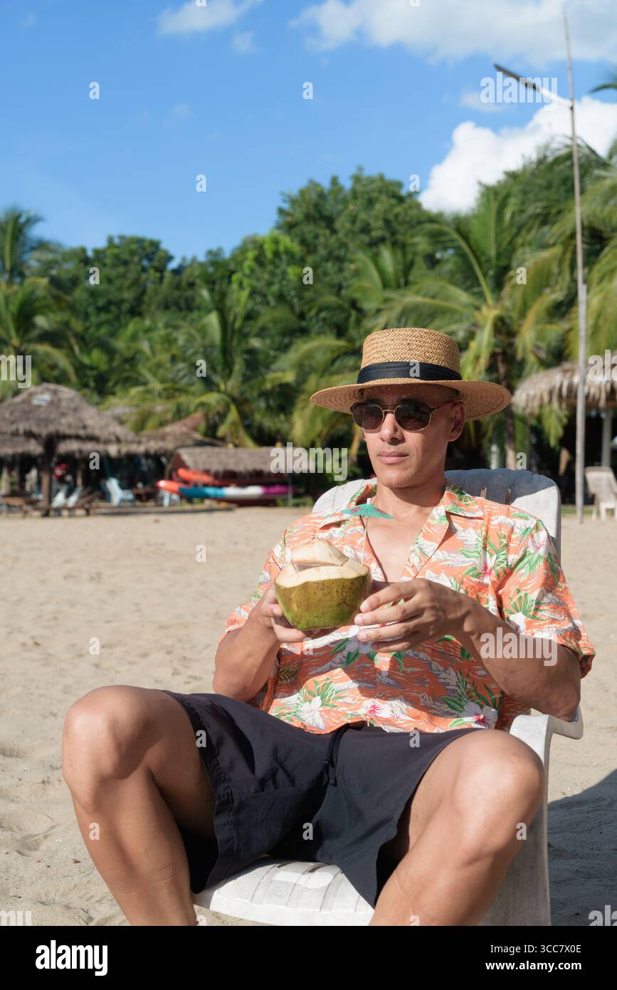 Hispanischer Mann mit Panamahut und hawaiianischem Hemd, gekleidet im lässigen Sommerurlaub-Stil. Er posiert am Strand und zeigt eine entspannte Urlaubsatmosphäre Stockfoto