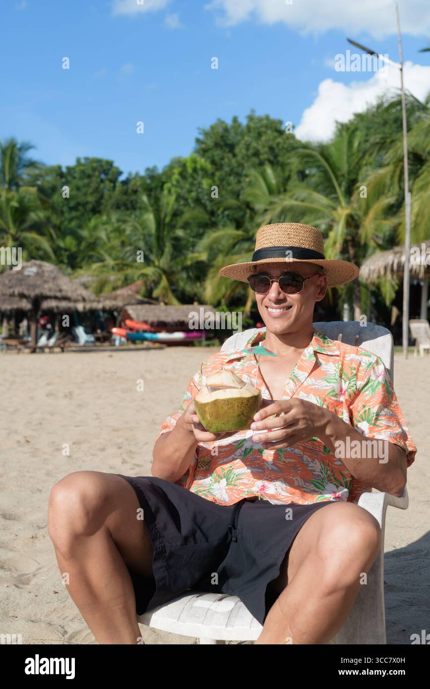 Hispanischer Mann mit Panamahut und hawaiianischem Hemd, gekleidet im lässigen Sommerurlaub-Stil. Er posiert am Strand und zeigt eine entspannte Urlaubsatmosphäre Stockfoto