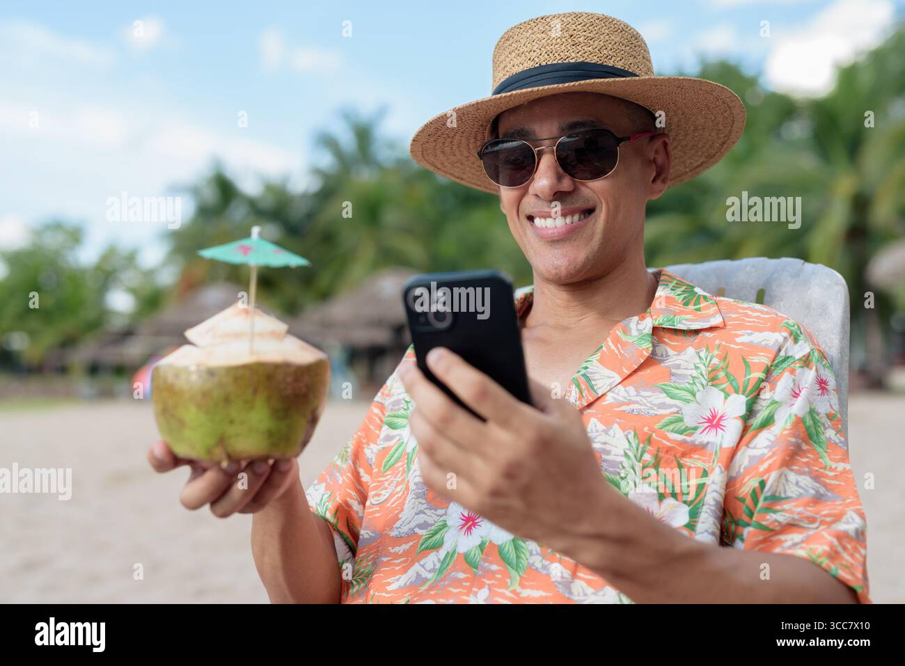 Hispanischer Mann mit Panamahut und hawaiianischem Hemd, gekleidet im lässigen Sommerurlaub-Stil. Er posiert am Strand und zeigt eine entspannte Urlaubsatmosphäre Stockfoto
