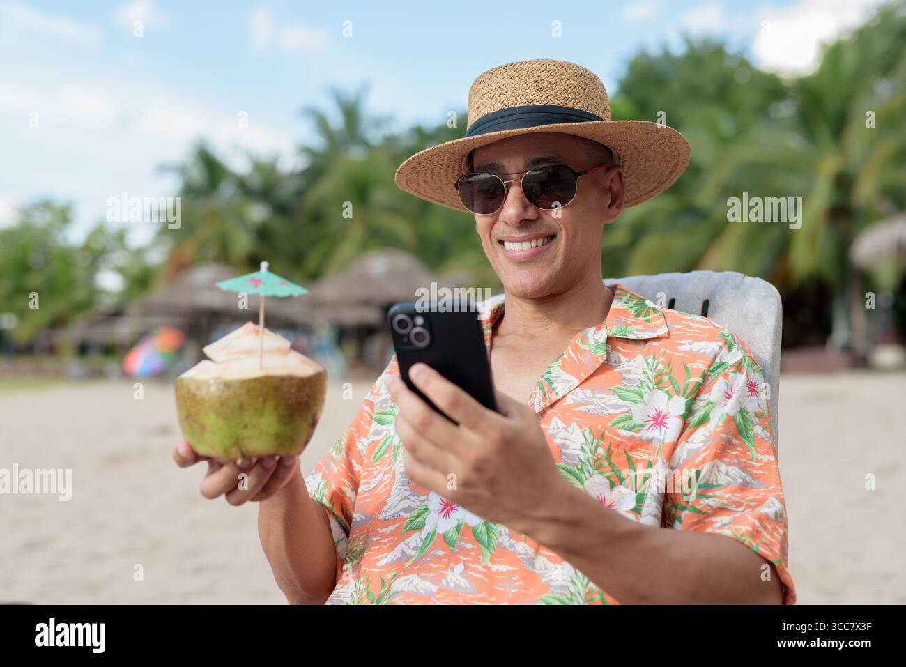 Hispanischer Mann mit Panamahut und hawaiianischem Hemd, gekleidet im lässigen Sommerurlaub-Stil. Er posiert am Strand und zeigt eine entspannte Urlaubsatmosphäre Stockfoto