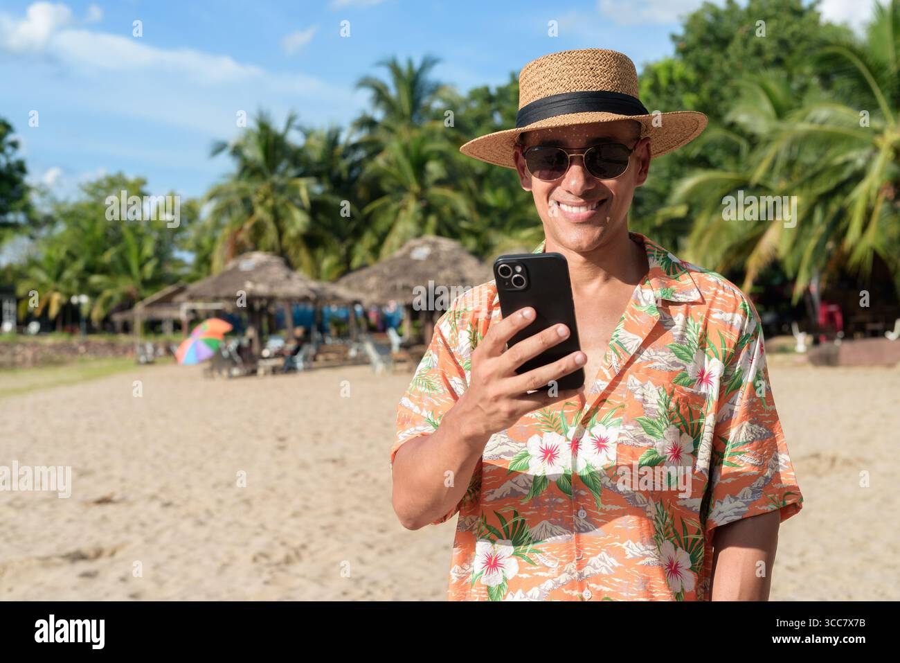 Hispanischer Mann mit Panamahut und hawaiianischem Hemd, gekleidet im lässigen Sommerurlaub-Stil. Er posiert am Strand und zeigt eine entspannte Urlaubsatmosphäre Stockfoto