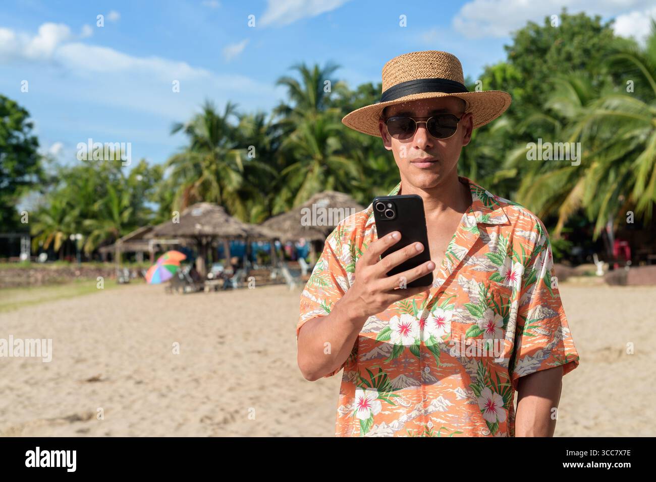 Hispanischer Mann mit Panamahut und hawaiianischem Hemd, gekleidet im lässigen Sommerurlaub-Stil. Er posiert am Strand und zeigt eine entspannte Urlaubsatmosphäre Stockfoto