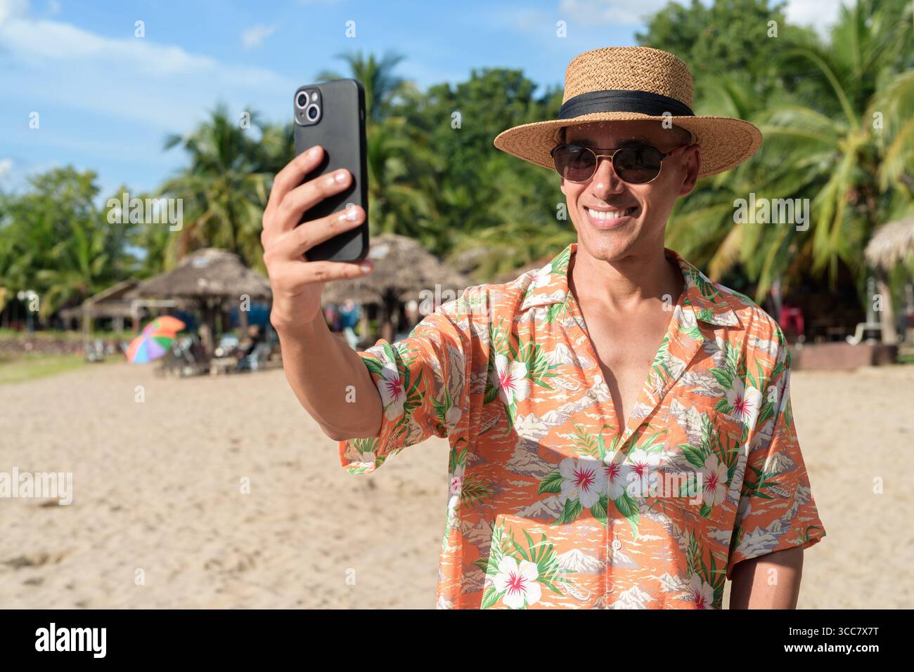 Hispanischer Mann mit Panamahut und hawaiianischem Hemd, gekleidet im lässigen Sommerurlaub-Stil. Er posiert am Strand und zeigt eine entspannte Urlaubsatmosphäre Stockfoto