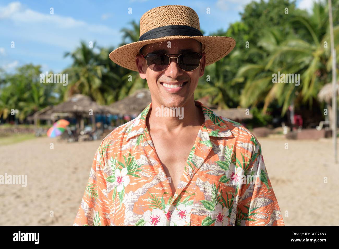 Hispanischer Mann mit Panamahut und hawaiianischem Hemd, gekleidet im lässigen Sommerurlaub-Stil. Er posiert am Strand und zeigt eine entspannte Urlaubsatmosphäre Stockfoto