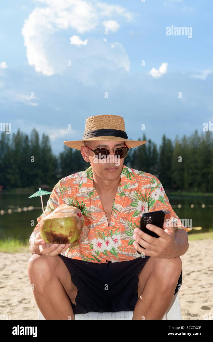 Hispanischer Mann mit Panamahut und hawaiianischem Hemd, gekleidet im lässigen Sommerurlaub-Stil. Er posiert am Strand und zeigt eine entspannte Urlaubsatmosphäre Stockfoto