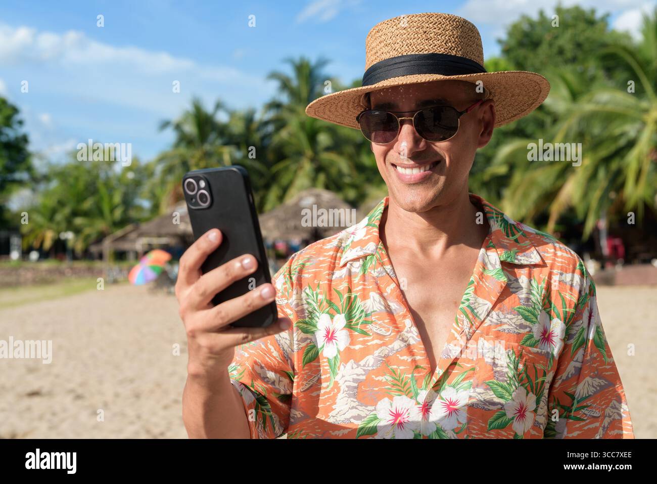 Hispanischer Mann mit Panamahut und hawaiianischem Hemd, gekleidet im lässigen Sommerurlaub-Stil. Er posiert am Strand und zeigt eine entspannte Urlaubsatmosphäre Stockfoto