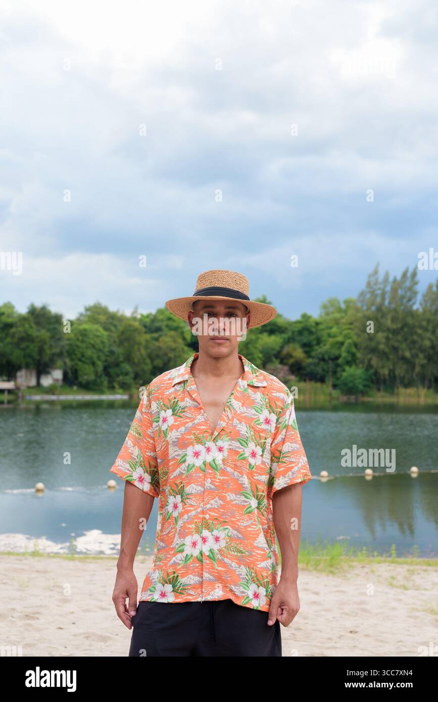 Hispanischer Mann mit Panamahut und hawaiianischem Hemd, gekleidet im lässigen Sommerurlaub-Stil. Er posiert am Strand und zeigt eine entspannte Urlaubsatmosphäre Stockfoto