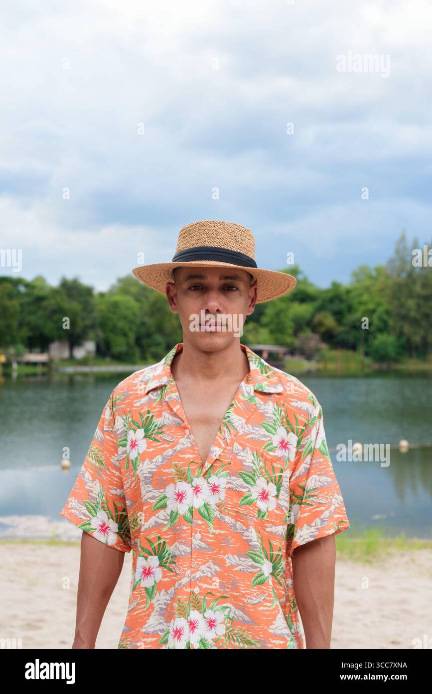 Hispanischer Mann mit Panamahut und hawaiianischem Hemd, gekleidet im lässigen Sommerurlaub-Stil. Er posiert am Strand und zeigt eine entspannte Urlaubsatmosphäre Stockfoto