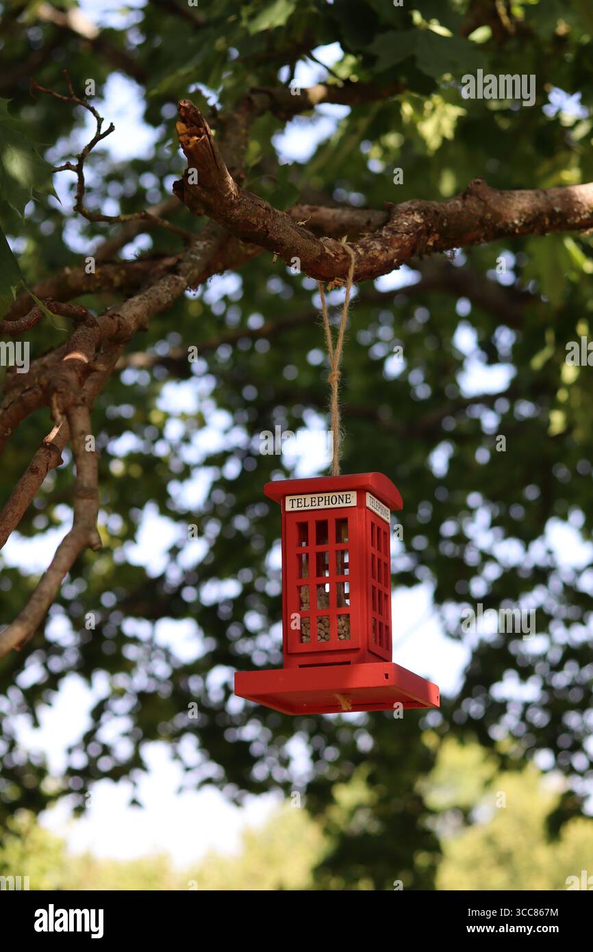 Ein charmanter Vogelfutterplatz in Form einer klassischen roten britischen Telefonzelle hängt an einem Baumzweig in einem sonnigen Park. Stockfoto Ein charmanter Vogelfutterplatz in Form einer klassischen roten britischen Telefonzelle hängt an einem Baumzweig in einem sonnigen Park. Stockfoto