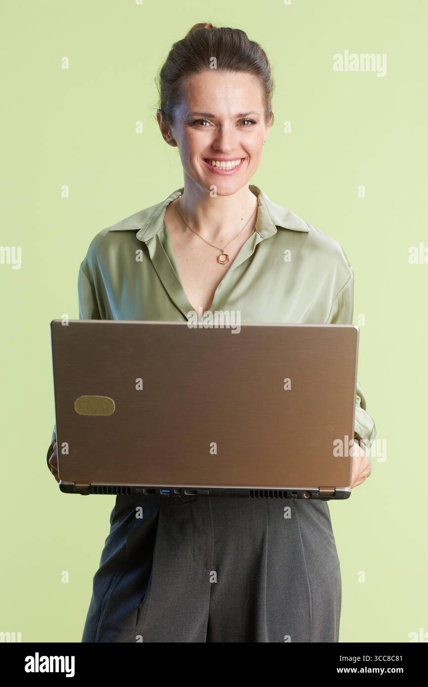 Eine lächelnde Frau im Alter von 30 Jahren hält einen Laptop und strahlt Vertrauen und Zugänglichkeit vor einem weichen, hellgrünen Hintergrund in einem professionellen Studio aus Stockfoto