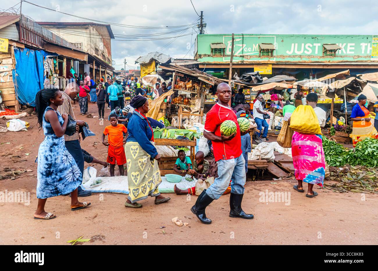 Ein einheimischer Afrikaner trägt zwei große Melonen auf einem geschäftigen Lebensmittelmarkt in einem Dorf in der Nähe von Kampala, Central Region, Uganda Stockfoto