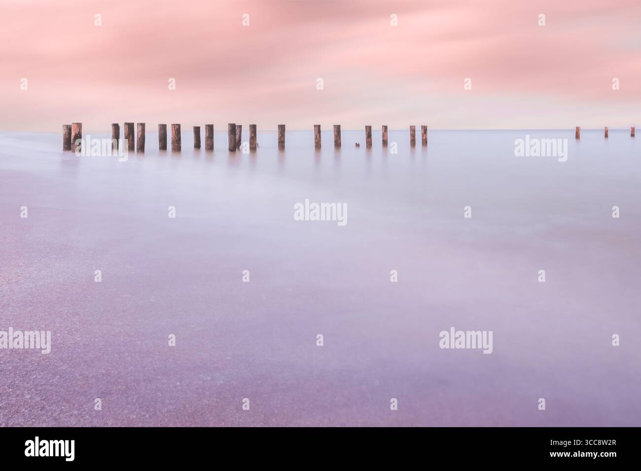 Eine traumhafte Meereslandschaft mit einem pastellfarbenen Himmel, der sich auf ein ruhiges Meer reflektiert, mit Holzpfosten, die im sanften Wasser stehen und Ruhe und Ruhe wecken Stockfoto