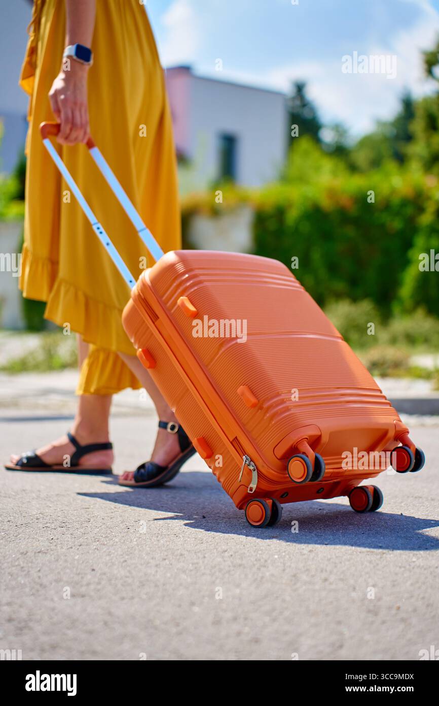 Frau in gelbem Kleid mit orangem Koffer, die an sonnigem Tag auf der Straße in der Nähe des Wohnhauses läuft. Weibliche Reisende mit Gepäck kehren von der Reise nach Hause zurück Stockfoto