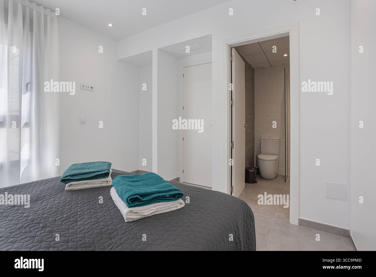 Minimalistisches Schlafzimmer mit einem ordentlich gefertigten Bett mit gefalteten Handtüchern, direktem Zugang zu einem modernen Bad und hellem Tageslicht Stockfoto