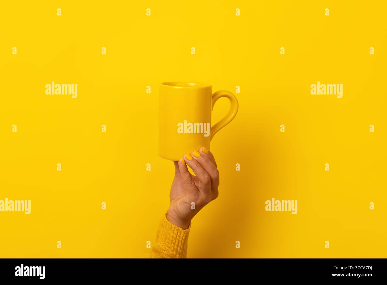 Hand mit gelben Nägeln und Pullover, der einen einfarbigen gelben Becher vor einem nahtlosen gelben Hintergrund hält, sorgt für einen monochromatischen Look Stockfoto
