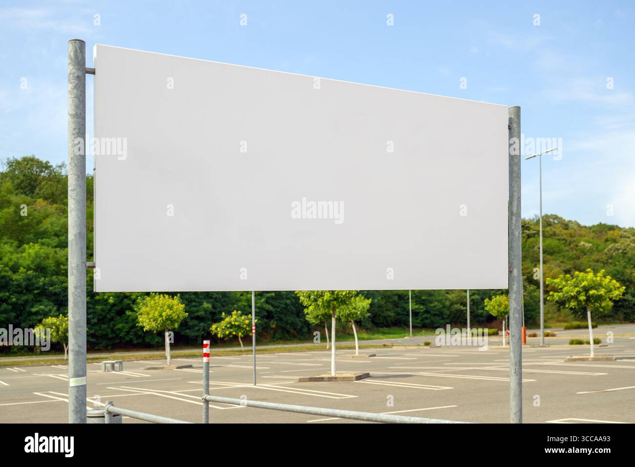 Leere weiße Mockup-Werbetafel auf einem leeren Außenparkplatz mit Bäumen im Hintergrund an einem sonnigen Tag. Selektiver Fokus. Stockfoto