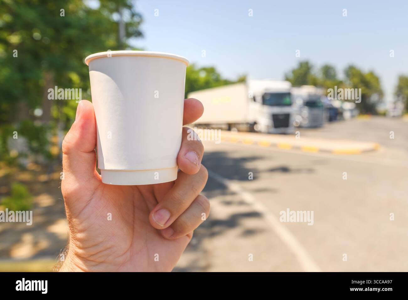 Hand hält eine leere Kaffeetasse aus Papier an einer LKW-Haltestelle mit geparkten Aufliegern im Hintergrund. Selektiver Fokus. Stockfoto