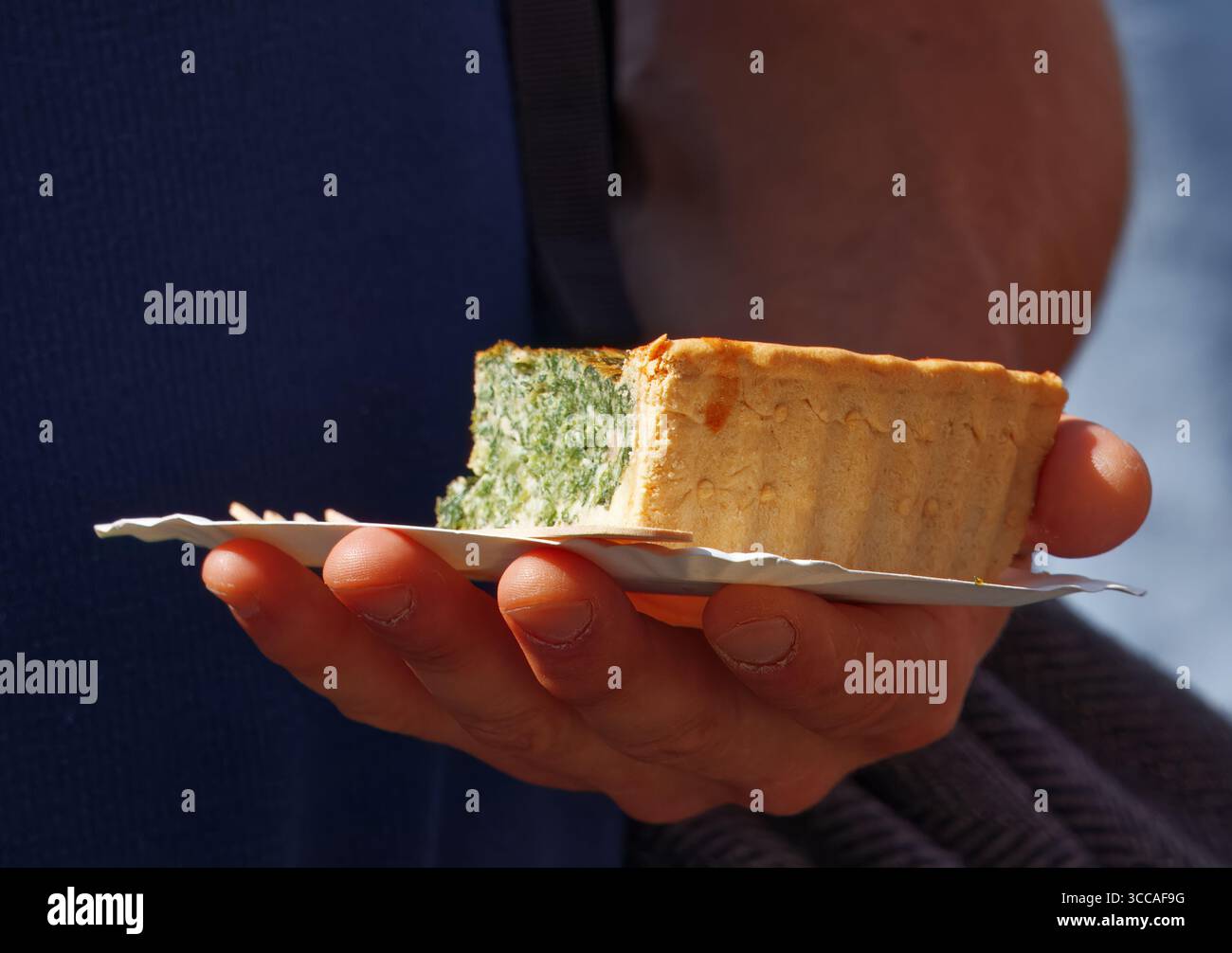 Nahaufnahme einer Hand, die ein Stück Spinatquiche auf einem Papierteller hält Stockfoto