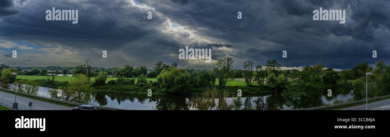 Dunkle Wolken schweben über die Donau in Niederbayern, während das schwindende Licht des Tages die Landschaft erleuchtet. Grüne Wiesen und Bäume umrahmen die ruhige Wa Stockfoto