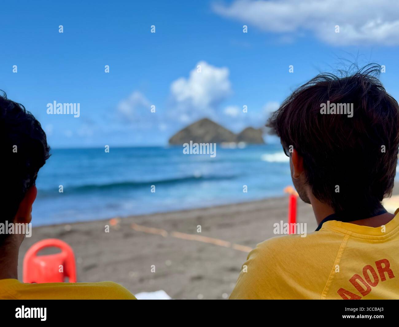 Zwei Rettungsschwimmer beobachten das Meer von einem Außenposten am Strand von Mosteiros, Azoren, Portugal, mit vulkanischen Inseln in der Ferne. - Smartphone-aufgenommenes Stockfoto