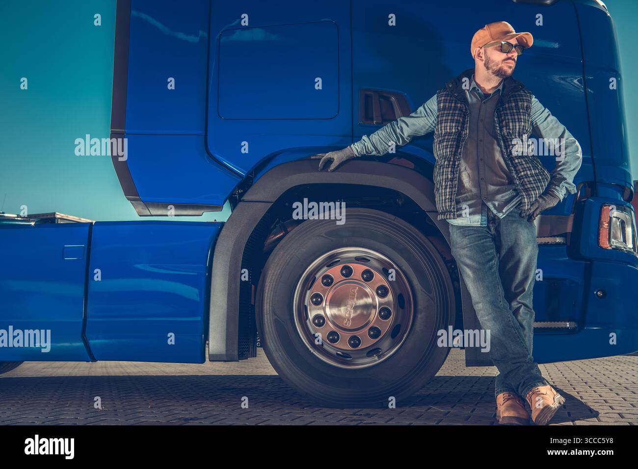 Ein Lkw-Fahrer lehnt sich an ein blaues LKW-Rad und genießt den sonnigen Tag. Er trägt ein lässiges Outfit mit einer Sonnenbrille und einer Mütze, die einen entspannten AT projiziert Stockfoto