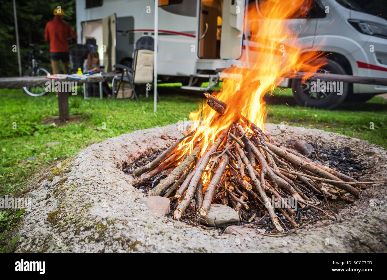 Freunde genießen im Sommer ein gemütliches Lagerfeuer auf einem üppigen Campingplatz, umgeben von Bäumen und einem geparkten Wohnmobil, was eine warme und einladende Atmosphäre schafft. Stockfoto