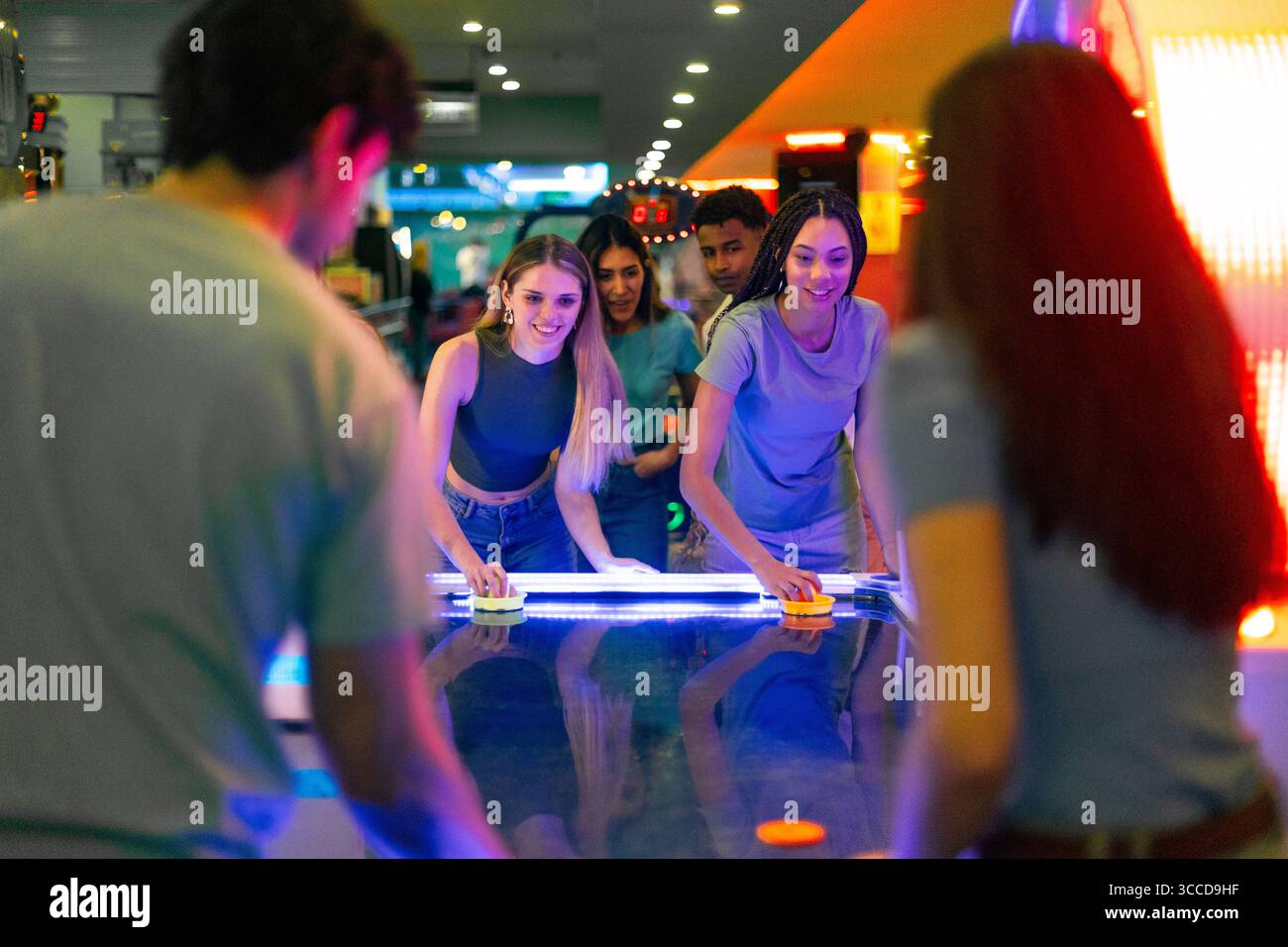 Eine Gruppe von Freunden genießt aufregende Momente beim Spielen von Air Hockey in einem lebhaften Arcade-Spielraum voller farbenfroher Lichter Stockfoto