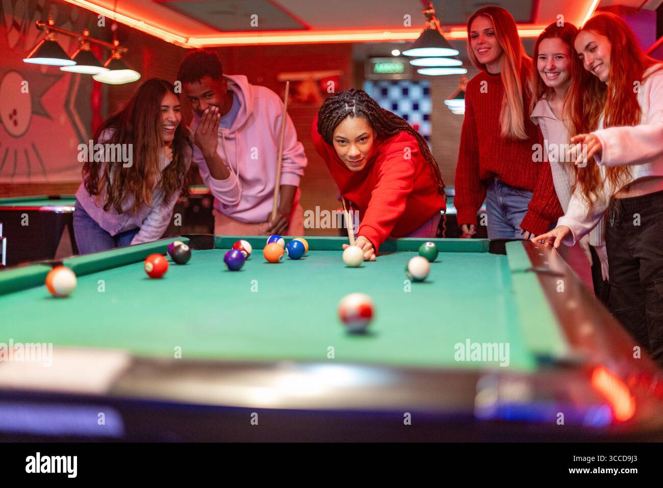 Die junge Frau schießt im Billard, während ihre Freunde sie in einer pulsierenden Billardhalle beobachten und anfeuern Stockfoto