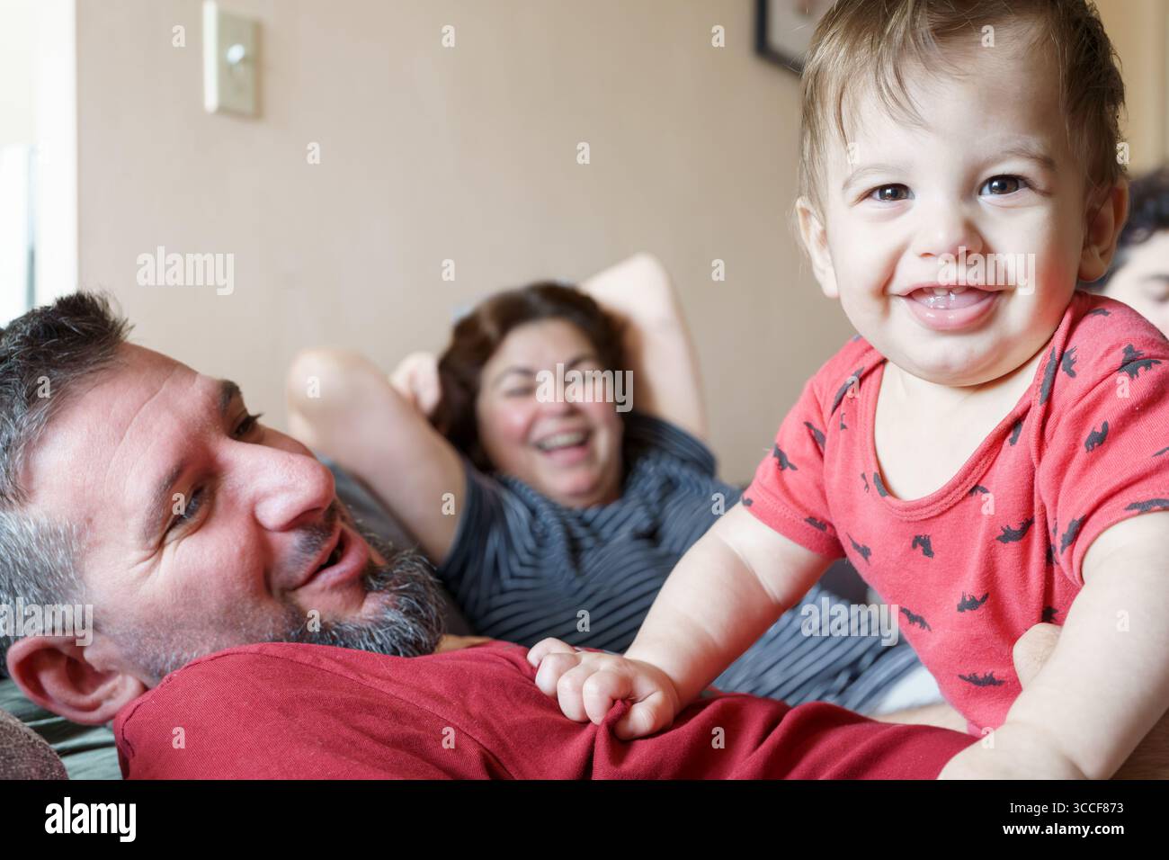Lächelndes Baby in roter Kleidung beugt sich über den Mann, während die Frau im Hintergrund lacht und einen fröhlichen Familienmoment in Innenräumen festhält. Stockfoto
