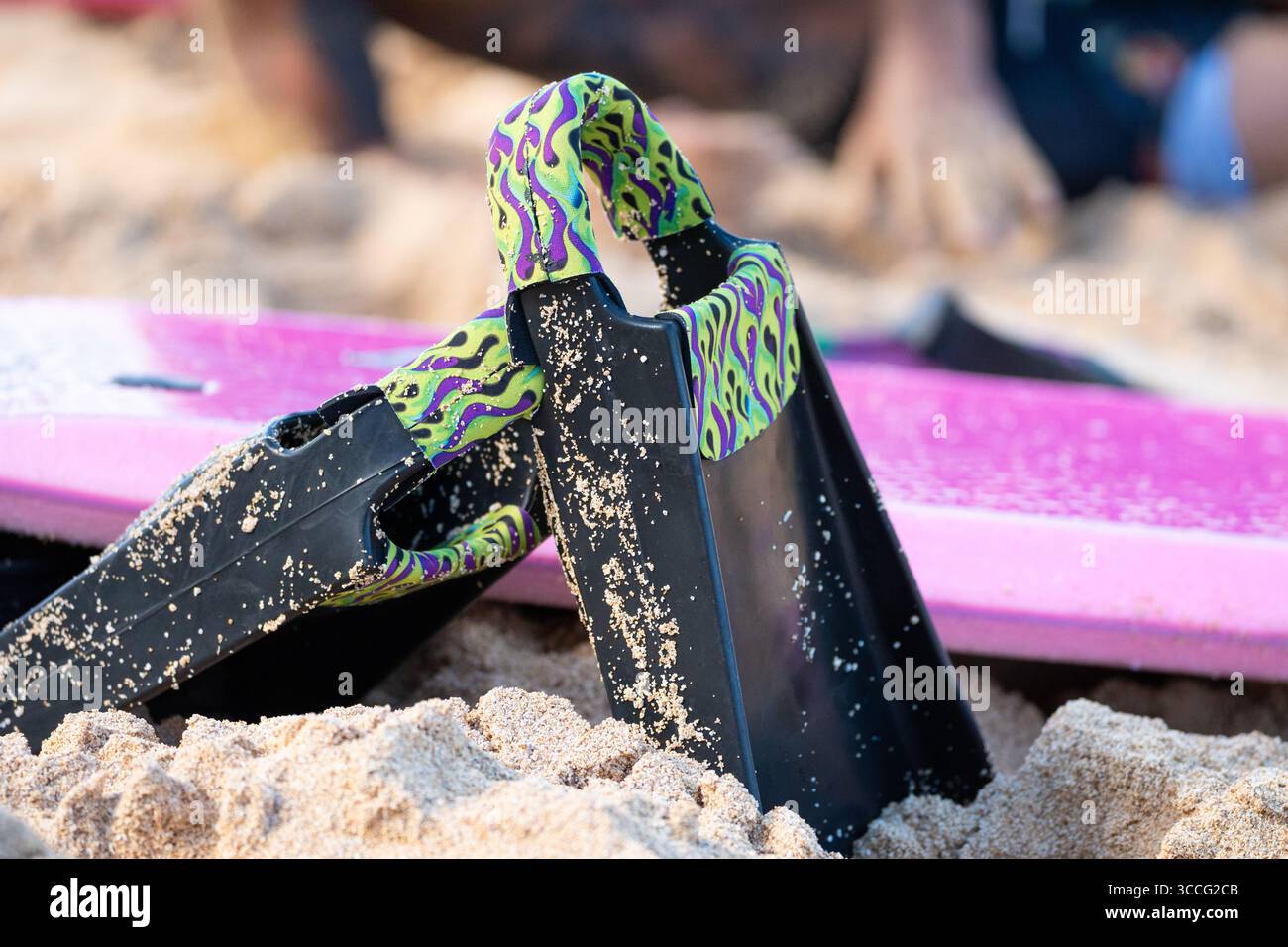 Zwei schwarze Schwimmflossen mit violettem und grünem Flammendesign, die auf goldenem Sand ruhen, mit passendem lila Bodyboard im Hintergrund unter dem sonnigen Strandhimmel Stockfoto