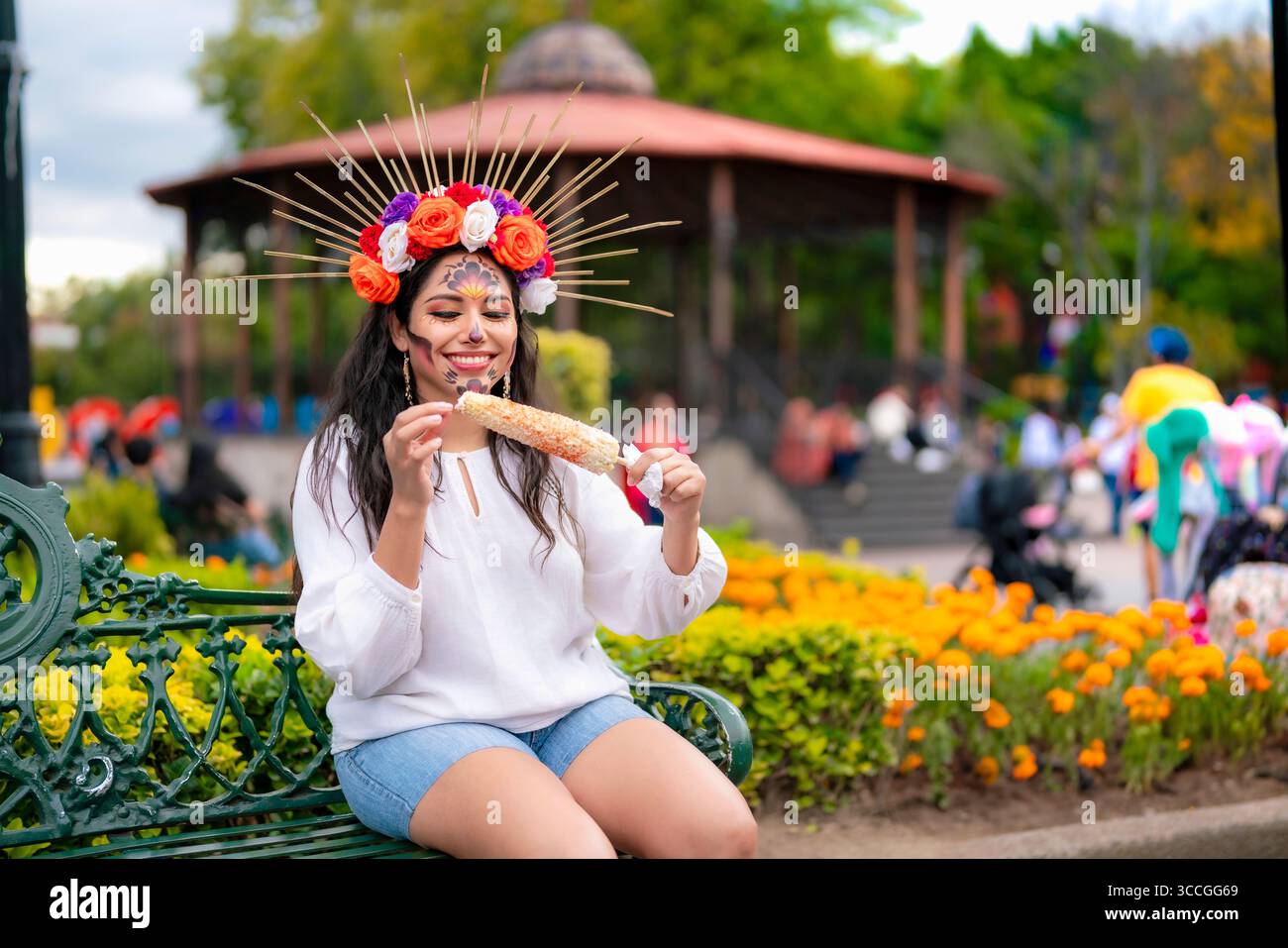 Latina-Frau mit catrina-Make-up und Blumenkrone isst eine traditionelle Elote, während sie im Coyoacan Park sitzt, umgeben von Cempasuchil-Blumen und einem kios Stockfoto