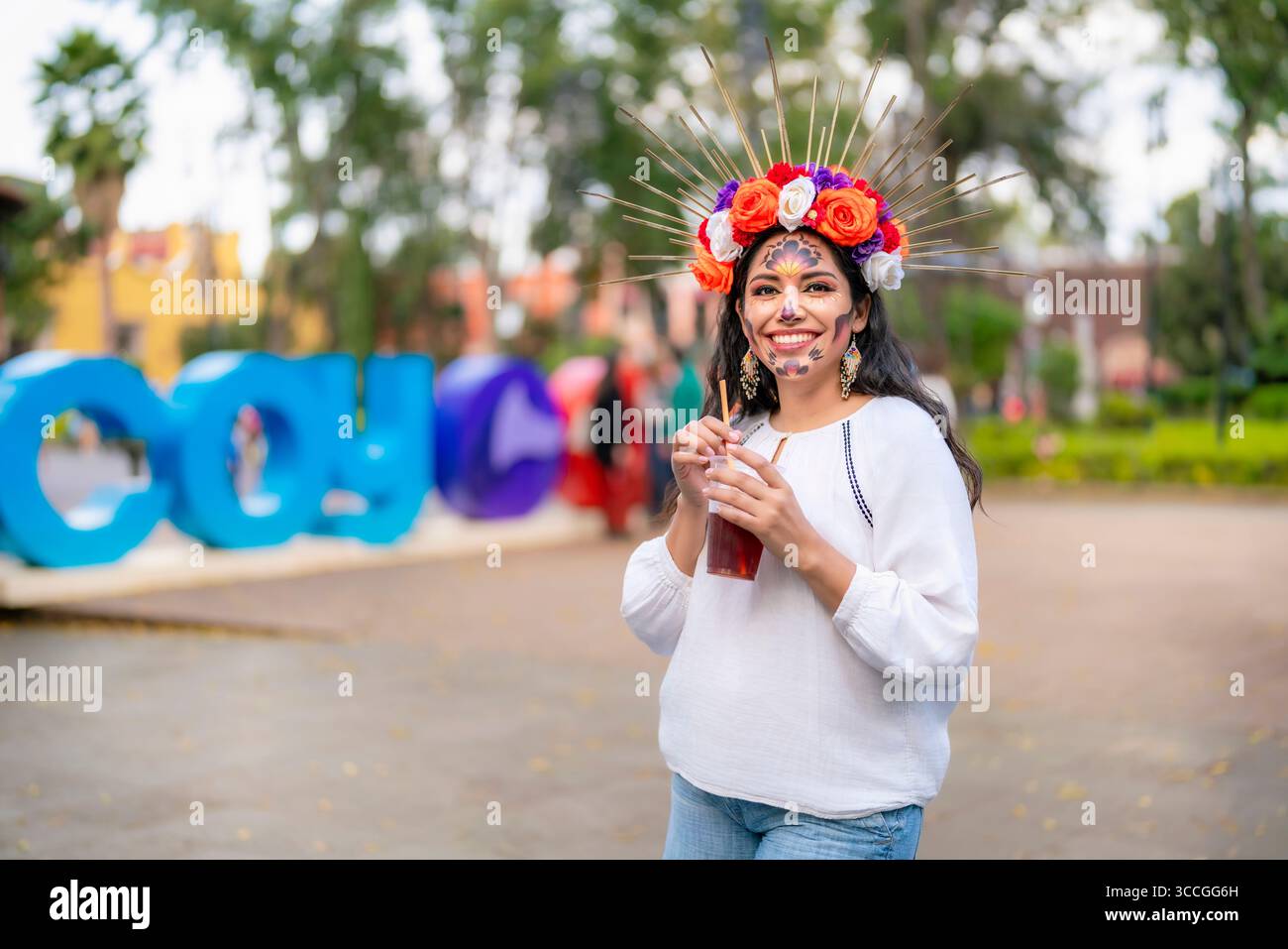Die junge Latina mit catrina Make-up und farbenfroher Blumenkrone lächelt beim Trinken eines traditionellen mexikanischen Getränks im Coyoacan Park, feiert Dia Stockfoto