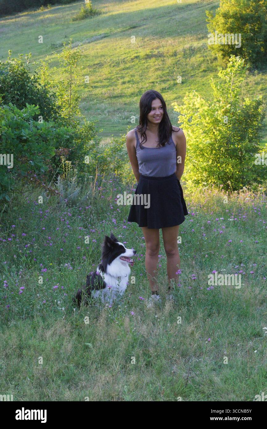 Frau, die mit Border Collie neben ihr auf einer grasbewachsenen Wiese steht, während der sonnigen Sommertage. Ideal für Gehorsam bei Haustieren, Trainingsroutinen und Outdoor Stockfoto