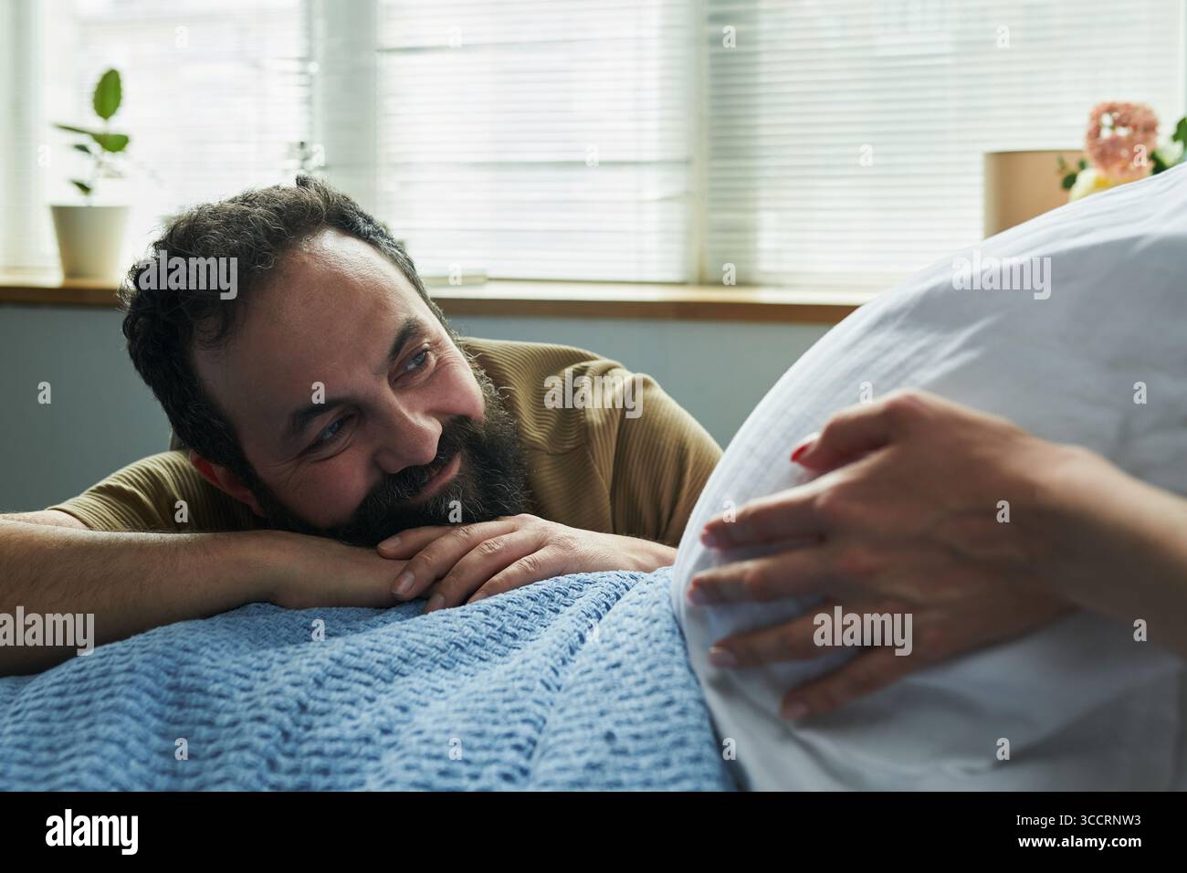 Kaukasischer Mann mittleren Alters lächelt und ruht Kopf auf den Händen, während er schwangere Frau ansieht, die den Bauch berührt, und zeigt einen liebevollen Moment zwischen werdenden Ehepaaren drinnen Stockfoto